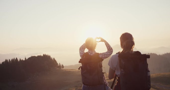 Friends on a hike in the mountains hiking together in early fall or autumn morning sunrise take photo with smartphone of hands love heart shape over the sun and smile happiness