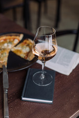 Close up of glass of rose wine with 4 cheese pizza and fresh newspaper on background at the cafe. Meal outside home. Leisure concept.