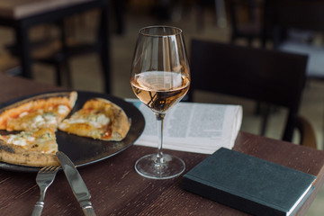 Glass of rose wine, 4 cheese pizza, newspaper and notebook on wooden table at the cafe. Day off meal outside home.