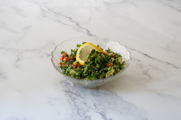A 45 degree angle of tabbouleh garnished with parsley and a lemon slice. Set against a marble white background with copy space available