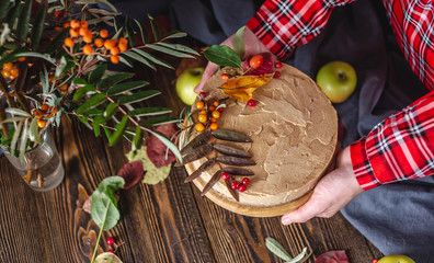 Women's hands are holding a mousse brown cake decorated with autumn leaves and berries. Concept of autumn atmosphere and mood