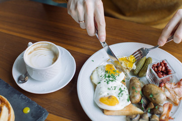 Close up of man's hands holding fork and knife while eating traditional English breakfast with fried eggs, sausages, beans, mushrooms, bacon.