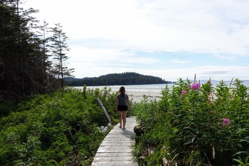 A female hiker walking along the boardwalk to nels bight surrounded by forest and the pacific ocean, along the beautiful cape scott trail on Northern Vancouver island