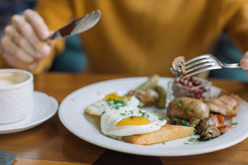Man eating traditional English breakfast with fried eggs, sausages, beans, mushrooms, bacon at cafe in the morning.