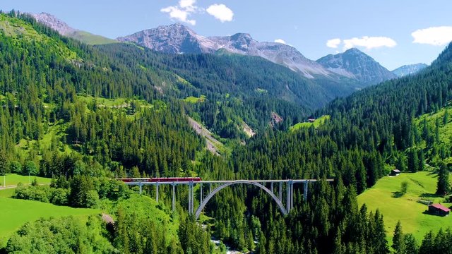 Train running on a bridge in the middle on a beautiful landscape. We can see pine forest and mountains in the background, under the summer sunlight - aerial view with a drone 4K