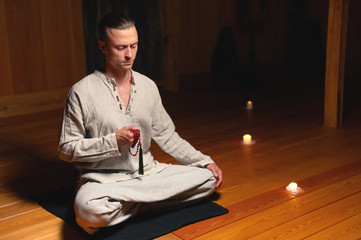 A portrait of an attractive Caucasian man sits in cotton robes in a lotus pose in a dark wooden...