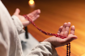 close-up man in clothes for practice and meditation sits in a lotus pose and holds red rosary to concentrate attention in a wooden room with dim light