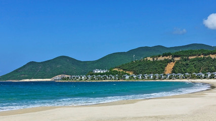 Tropical beach. Curved coastline, turquoise waves with white foam run on the white sand. There are a number of villas and palm trees in the distance. Green mountains against the blue sky. Vietnam.