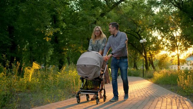 A happy heterosexual couple walks in the Park with a baby stroller. A young wife and her husband are walking in the Park holding hands on the stroller and smiling talking