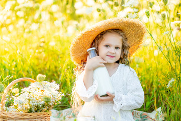 a beautiful little girl is sitting in a straw hat in a yellow field with wild flowers and holding a bottle of milk