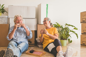 Smiling couple of senior people white hair working in the new empty apartment with moving boxes, sitting on the floor having break - concept of active elderly people and new beginning like retired