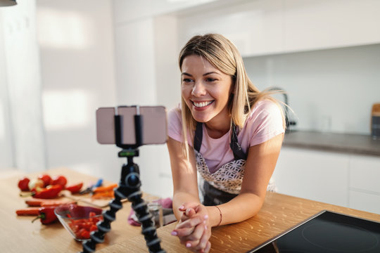 Young smiling gorgeous blond housewife leaning on kitchen table and looking at smart phone on tripod. She is looking at recipe for healthy lunch.
