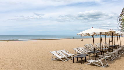 Beach umbrella and deck chairs at foreground. Scenic view of empty tropical sandy and peaceful beach under cloudy blue sky with light turquoise waves. 
