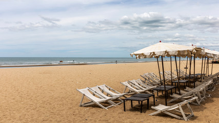 Beach umbrella and deck chairs at foreground. Scenic view of empty tropical sandy and peaceful beach under cloudy blue sky with light turquoise waves. 