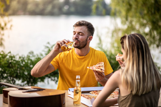 A young couple is sitting at a table in the garden, eating pizza and drinking beer in the background you can see the river