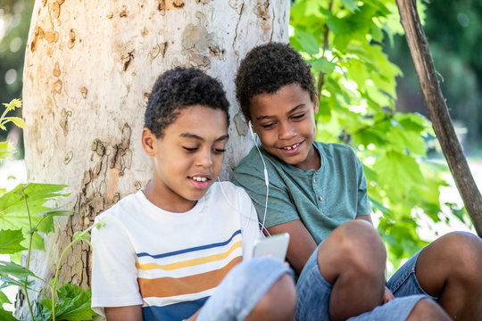 Two boys are sitting under a tree in the shade and listening to music