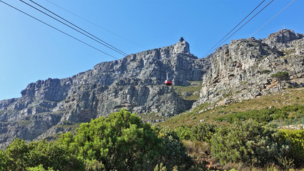 Fototapeta na wymiar Cable way to Table Mountain, Cape Town. The red round booth is lifted on ropes. Sheer cliff against a bright blue sky, green vegetation below. South Africa.