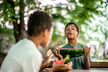 Two boys are sitting at a table, eating a watermelon and one is spitting at the other with a watermelon seed