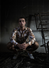 Young male in a white print shirt crouching in an abandoned building