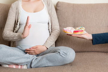 Close up of pregnant woman sitting on the sofa rejects to eat junk food such as donuts and makes no gesture. Healthy diet for future mother concept