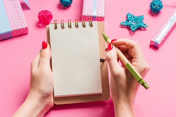 Top view of female hand making some notes in noteebok on pink background. New Year decorations and toys. Christmas time concept