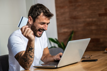 The man holds the card in his hand and prefers to look at the laptop after he has just done his shopping