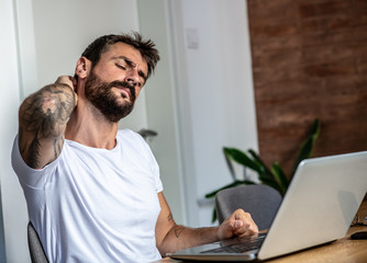 A male freelancer is looking at his laptop and holding his neck, he has neck pain