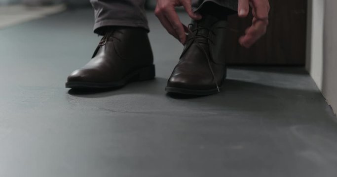 Slow motion dolly shot of young man unlace his brown leather chukka boots indoor