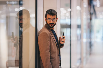 Man stands leaning against a shop window, holding a cell phone in his hand and looking at the store