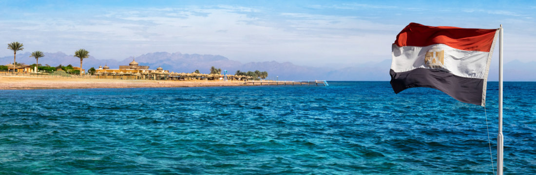 Aqaba bay view near the Dahab resort with a hotel, palms and silhouettes of mountains at skyline. Panorama of the coastline of Red Sea with Egyptian flag on a beautiful beach in Egypt.