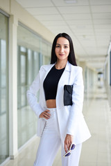 Young brunette woman, wearing white pants and jacket, standing in light passageway with huge windows, holding black clutch purse, posing for social media. Businesswoman on a break. Female portrait.