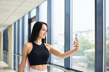 Fototapeta na wymiar Young brunette woman, wearing white pants and black top, holding jacket in hand, taking selfie picture with cell phone in light passageway, posing for social media. Businesswoman on lunch break.