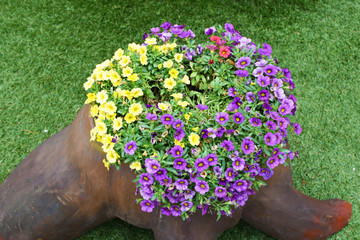 Colorful violet and yellow flowers in wooden pot made of tree trunk. Flowerbed with blooming flowers in retro planters on green artificial grass cover outdoors in sunny summer day.
