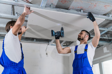 A builder and his colleague fix a plasterboard plate to the ceiling
