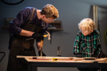 a young male carpenter is teaching woodwork to a young boy in his workshop.