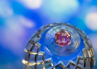 Close up of a pink transparent d20 polyhedral dice on top of a crystal dome against a blurry background.