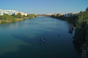 Fototapeta na wymiar Vistas del río Guadalquivir a su paso por la ciudad de Sevilla con personas haciendo paddle surf y remo. Al fondo se puede ver el puente de Isabel II más conocido como puente de Triana.