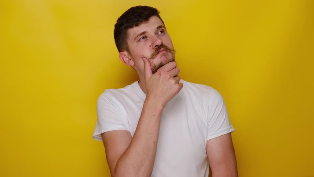Portrait of thoughtful confused young man touching chin, scratching head while thinking over solution, guy having doubts about difficult choice, wears t-shirt, isolated on yellow studio background