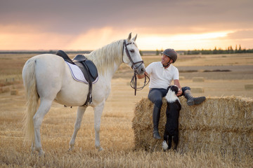 Man sitting on a straw bundle with his saddled horse and dog. Country scene.