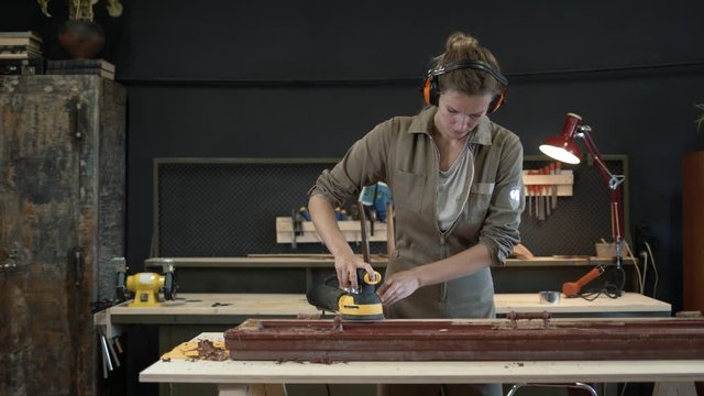 Young woman grinds the wooden door, restoration works. Female wearing hearing protection device grinding, polishing old door in workshop