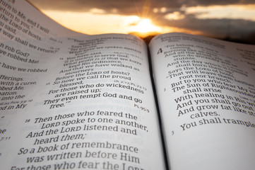 Holy Bible open at sunset with highlight on Malachi 4:2. Background with sun and clouds.