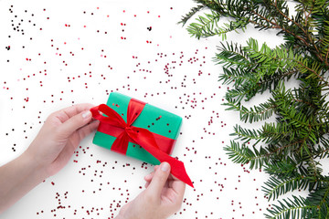 Hands hold one Christmas gift with red ribbon on the white table with Christmas tree nearby . Christmas wrapping