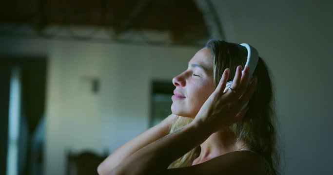 Authentic close up shot of young happy smiling woman is putting wireless headphones on and having fun to dance while listening her favorite music behind a window in a bedroom with a daylight at home.