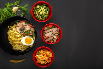 Noodles with pieces of meat and egg in a black plate and dim sum with different traditional snacks of mushrooms, soy and vegetables on a dark table. View from above. Asian food concept. Copy space