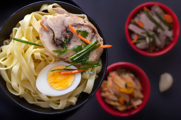 Noodles with pieces of meat and egg in a black plate and dim sum with different traditional snacks of mushrooms, soy and vegetables on a dark table. View from above. Asian food concept. Close-up