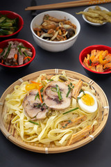 Noodles with chunks of meat and egg in a bamboo and dim sum plate with various traditional mushroom, soy and vegetable appetizers on a dark table. Asian food concept