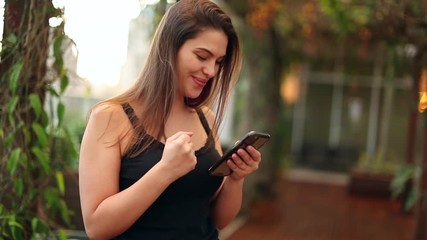 Happy young woman celebrates news holding smartphone. Girl doing positive gesture with fist