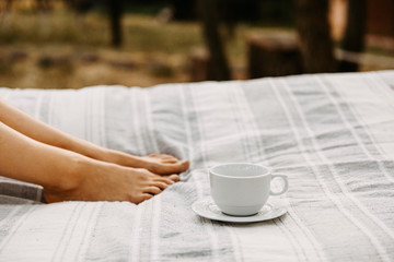 White blank cup of coffee on a bed with female legs on the background.