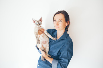 Obraz premium Lifestyle photo of a casual dressed female in blue jeans shirt holding and cuddling cute and funny Devon Rex cat. Happy kitty in girls arms. Breed with special hypoallergenic, no low shedding fur