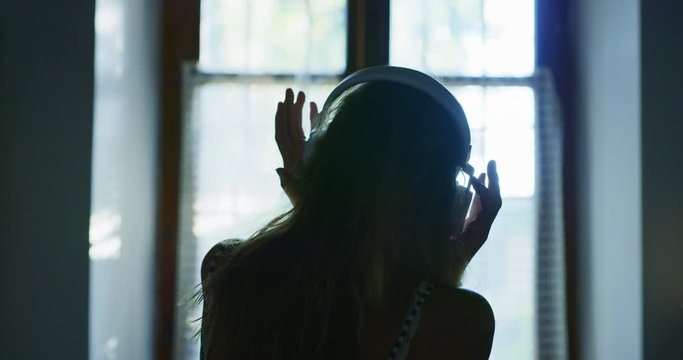 Authentic close up shot of young happy woman is putting wireless headphones on and having fun to dance while listening her favorite music behind a window in a bedroom with a daylight at home.